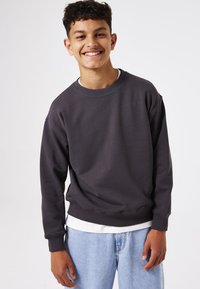 Sudadera gris oscuro con cuello redondo y puños acanalados, llevada sobre una camiseta blanca, combinada con unos vaqueros de color azul claro. Textura suave.