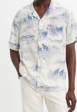 Homme portant une chemise à manches courtes boutonnée avec un imprimé paysage désertique bleu et cheval, et un pantalon blanc, mains dans les poches.
