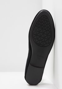 Semelle de chaussure plate noire avec un motif de bande de roulement géométrique et la taille 37 indiquée, positionnée à la verticale contre une surface d'angle blanche.