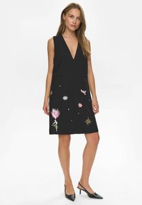 Vestido negro sin mangas con un profundo escote en V, dos bolsillos frontales y coloridos motivos bordados de flores y estrellas en la parte inferior delantera.