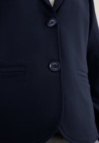 Marinblå blazer i sammetsliknande tyg, med två knappar och två sidofickor, samt slagkrage och en elegant design.