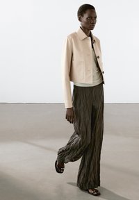 Beige cropped læderjakke med to frontlommer, parret med en lys grøn top og plisserede mørke grå brede bukser.