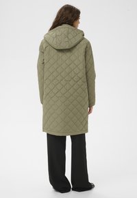 Abrigo largo acolchado verde oliva con capucha, con un patrón de rombos y dobladillo recto. Combinado con pantalones negros de pierna ancha.