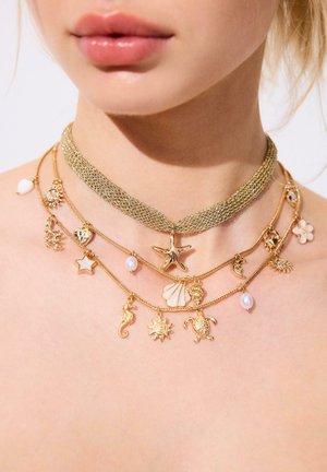 Geschichtete Goldketten mit ozeanisch inspirierten Anhängern, darunter ein Seestern, eine Muschel, eine Schildkröte, Sonne, Mond und Perlen, die den Hals und die Brust einer Frau schmücken.