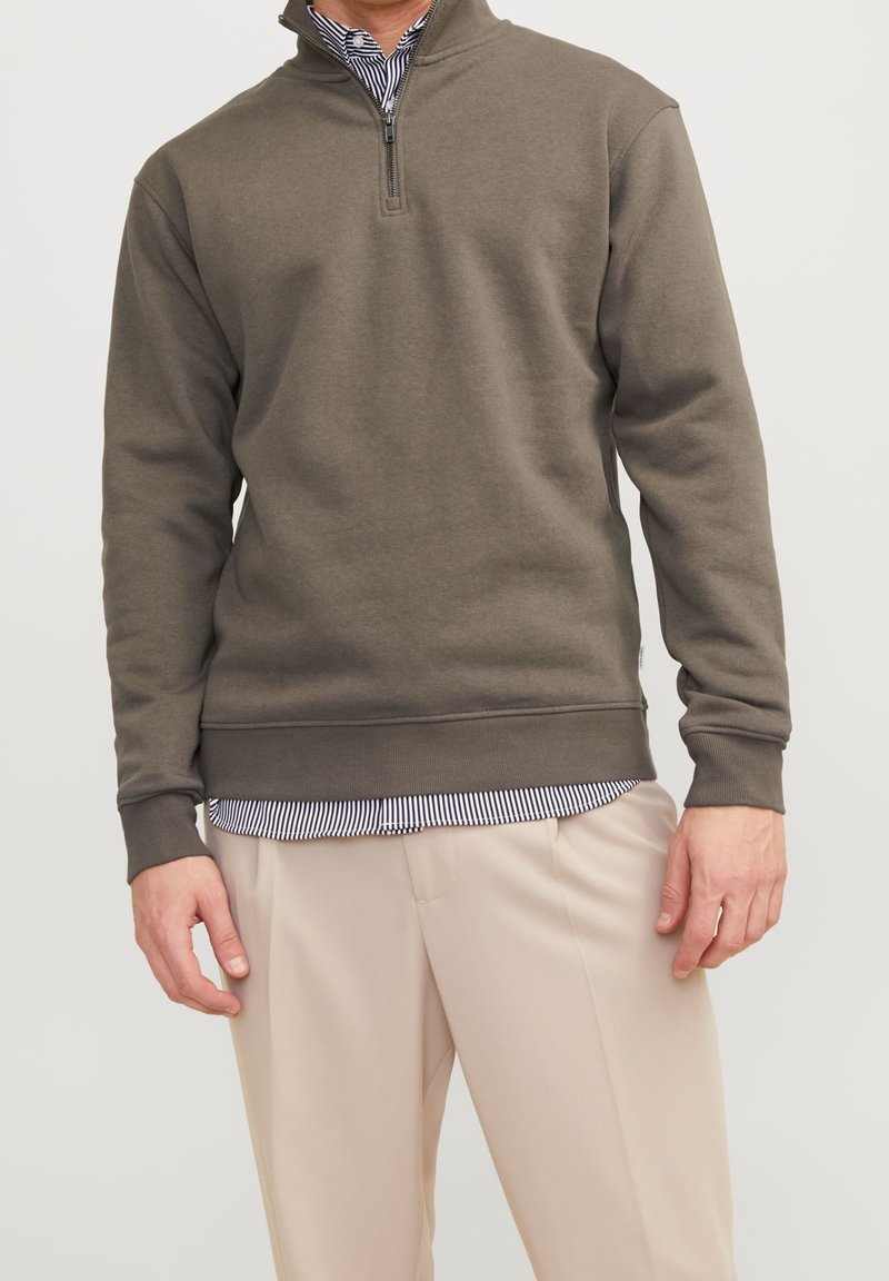 Pullover in verde oliva con zip a un quarto, maniche a costine e orlo a costine, abbinato a una camicia collare a righe visibile sotto.