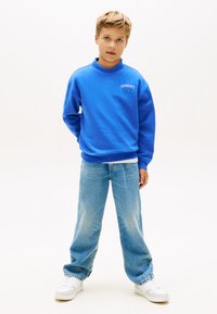 Μπλε βαμβακερό φούτερ με ψηλό γιακά και λεπτομέρεια λογότυπου, συνδυασμένο με τζιν παντελόνι σε ανοιχτό μπλε και λευκά αθλητικά παπούτσια.