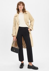Beige trenchcoat, crèmekleurig topje, op maat gemaakte zwarte broek en zwarte loafers. Zwarte handtas met gouden accenten en logo-details.