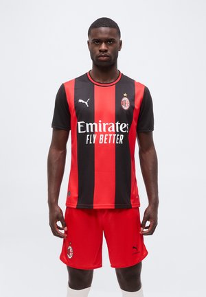 Muž v červeno-čiernom pruhovanom futbalovom drese AC Milan a červených šortkách, stojaci tvárou dopredu s neutrálnym výrazom.
