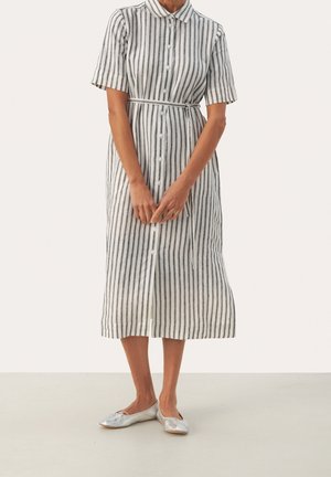 Femme portant une robe midi blanche à rayures noires verticales, manches courtes, col, fermeture à boutons sur le devant, ceinturée à la taille, et ballerines argentées.