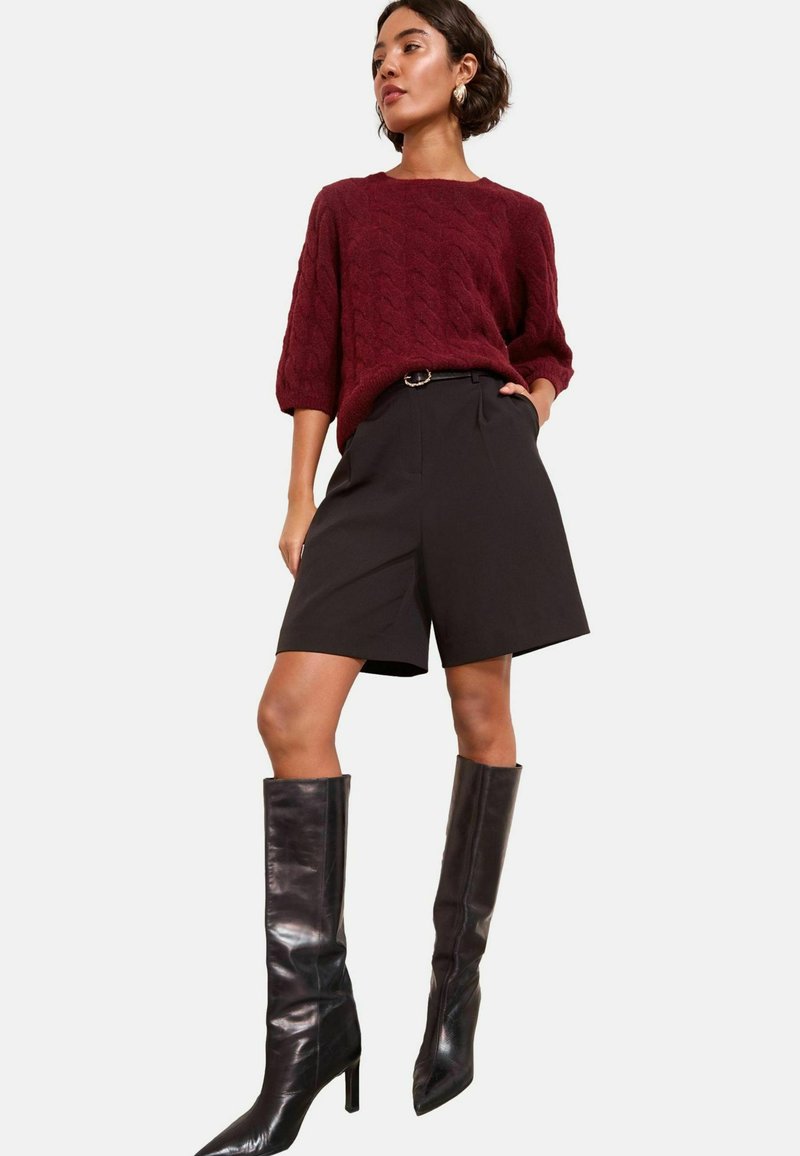 Burgundy, kötött pulóver háromnegyedes ujjakkal, fekete, testhez simuló sorttal és fekete, térdig érő, stiletto csizmával párosítva.