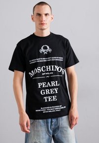 Juoda medvilnės marškinėliai su balta tekstu ir logotipais, trumpomis rankovėmis ir standartinio kirpimo. Išskirtinės detalės - prekės ženklas ir vieta.