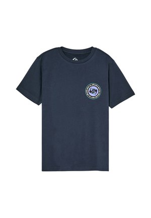 Dziecko z kręconymi włosami stojące z rękami za plecami, ubrane w granatowy t-shirt z małym okrągłym logo i luźne beżowe spodnie.