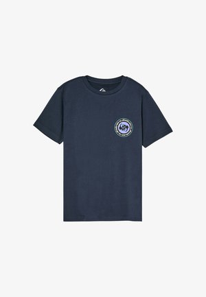 Dziecko z kręconymi włosami stojące z rękami za plecami, ubrane w granatowy t-shirt z małym okrągłym logo i luźne beżowe spodnie.