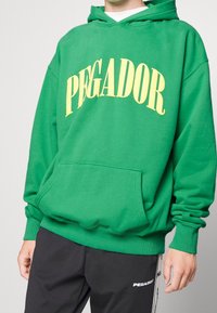 Persona che indossa un vivace hoodie verde con la scritta "PEGADOR" in giallo, abbinato a pantaloni neri con un logo sul lato.