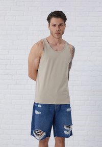 Beige tanktop med rund hals, parret med slidte blå denimshorts med frynser og flere huller. Modellen står op ad en hvid murstensvæg.