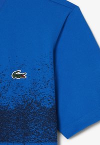 Camisa polo azul de manga corta, con una superficie texturizada, diseño en degradado negro y logotipo de cocodrilo verde bordado en la parte delantera.