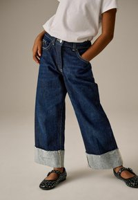 Jeans in denim blu a gamba larga con orlo sfilacciato di un blu chiaro, dotati di tasche frontali e cuciture a contrasto. Abbinati a scarpe nere con borchie.