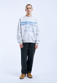 Gray sweatshirt with blue graphic text, relaxed fit, ribbed cuffs, and hem. Paired with black pants and tan sneakers.
