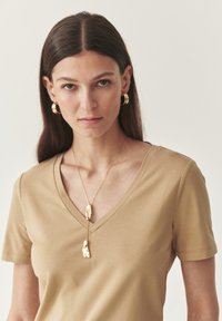 Beige v-ringad t-shirt tillsammans med guldörhängen i form av ringar och ett guldhalsband med oregelbundet formade hänge, mot en neutral bakgrund.