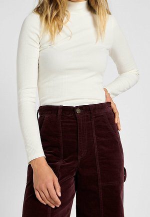 Personne portant un haut blanc côtelé à col roulé et manches longues, et un pantalon en velours côtelé violet foncé taille haute avec la main dans la poche.