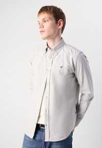 Lys grå skjorte med knapper laget av en bomullsblanding, med et lite logo på brystet, klassisk krage, og lange ermer. Brukt over jeans.