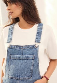 Pantalones vaqueros con tirantes azules ajustables, bolsillo frontal y herrajes plateados, llevados sobre una camiseta blanca de manga corta. Textura suave.