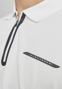 Hvid polo shirt med sort lynlås, kontrastkrave og detalje med sort mønstret logo på brystet. Struktureret stofdesign.