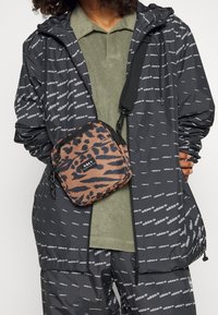 Černá větrovka s bílými potisky loga adidas, v kombinaci s olivově zeleným polo tričkem a hnědou kabelkou přes rameno s leopardím potiskem a nášivkou s logem.