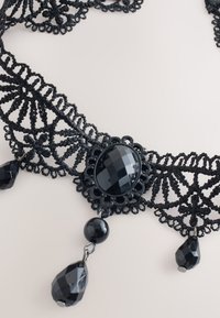 Black lace choker featuring an oval faceted pendant and two dangling beads. Intricate design with a decorative patterned edge.