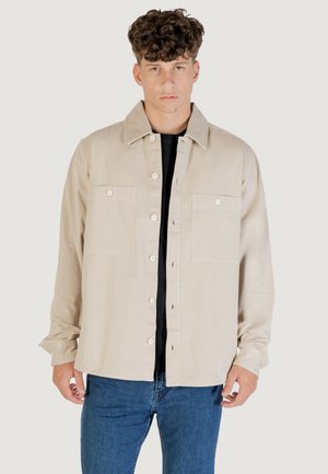 Young man with curly hair wearing a beige buttoned jacket over a black shirt and blue jeans, standing against a plain background.