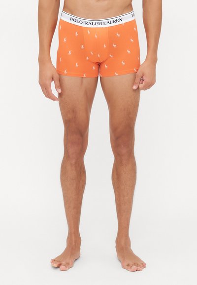 Moški nosi oranžne bokserice Polo Ralph Lauren z belim pasom z logotipom in majhnimi belimi motivi igralca polo na belem ozadju.