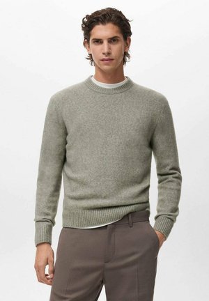 Joven con cabello rizado que lleva un suéter de cuello redondo gris claro sobre una camisa blanca y pantalones marrones, de pie con una mano en el bolsillo.