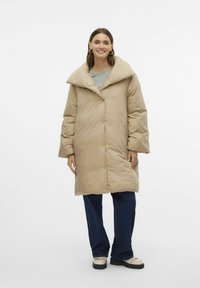 Beige puffy jakke med en stor krave, frontknapper og lange ærmer. Båret over en grå top, parret med mørke jeans og lyse sko.