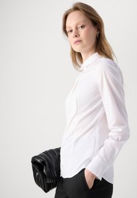 Bílá košile s límečkem a dlouhými rukávy, vyrobená z hladké tkaniny. V páru s černou skládací kabelkou.
