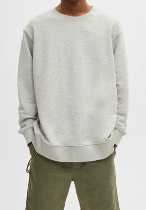 Person trägt ein hellgraues Sweatshirt mit Rundhalsausschnitt und olivgrüne Hose, die Hände in den Taschen, vor einem schlichten Hintergrund.