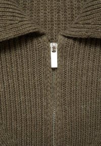 Hnědý pletený svetr s žebrovanou texturou, vybavený stříbrným zipem a překládaným límcem. Tkanina je zdá se hustá a teplá.