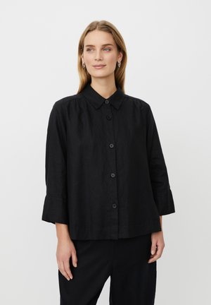 Frau mit langen blonden Haaren, die silberne Creolen und ein locker sitzendes schwarzes Hemd mit dreiviertel Ärmeln trägt, steht vor einem schlichten Hintergrund.