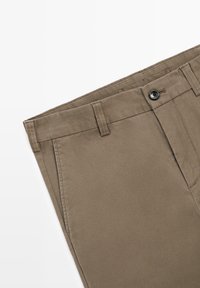 Khaki bukser med et glat, robust stof. Har knaplukning, bæltestropper og flad front, designet til afslappet eller semiformal påklædning.