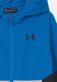 Blue athletic zip-up jacket with mesh hood, featuring a black logo on the chest and textured fabric. Includes a zipper closure.