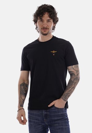 Uomo con capelli ricci che indossa una maglietta nera con piccolo emblema dorato e jeans blu, mostrando tatuaggi dettagliati su entrambe le braccia, con una mano in tasca.