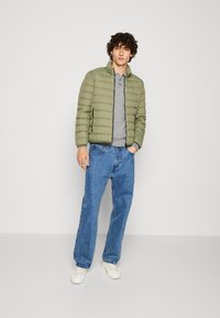 Giacca imbottita verde oliva chiaro con texture trapuntata, chiusura frontale a zip, indossata sopra una polo grigia, abbinata a jeans larghi blu e sneakers bianche.