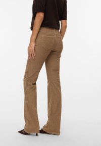 Femme portant un pantalon bootcut en velours côtelé marron, un haut noir et des chaussures à talons pointues, debout devant un fond uni.
