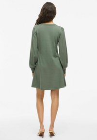 Abito lungo verde a maniche lunghe con scollatura rotonda, delicate pieghe sulle spalle e orlo svasato, realizzato in morbido tessuto testurizzato.