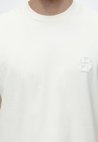 Άτομο που φοράει απλό λευκό T-shirt με στρογγυλή λαιμόκοψη και μικρό λευκό ανάγλυφο λογότυπο στην αριστερή πλευρά του στήθους.