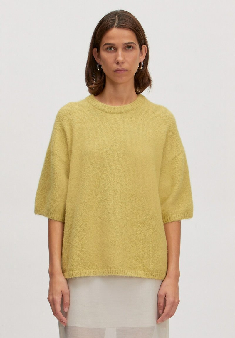 Gelber Strickpullover mit kurzen, weit geschnittenen Ärmeln, geripptem Ausschnitt und weichem Material. Über einem weißen durchsichtigen Kleidungsstück getragen.