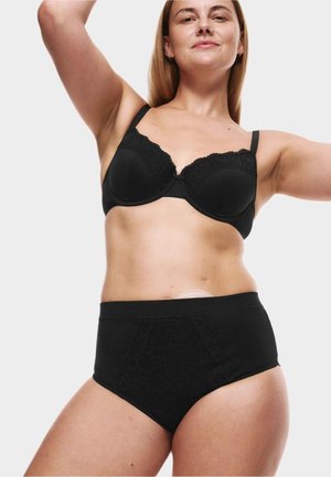 Kvinde iført sort blonder-bh og højtaljede trusser med synlig medicinsk stomi-pose på maven, armene løftet selvsikkert.