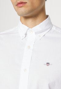 Hombre con camisa blanca abotonada con cuello abotonado y un pequeño logo bordado de GANT en rojo, blanco y azul en el pecho.