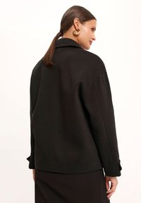 Czarna kurtka z wyrazistym fasonem, teksturowanym materiałem i zapięciem na guziki przy mankietach. Pokazana z tyłu, z uwydatnionym kołnierzem i bezszwowymi ramionami.
