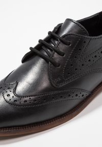 Černé kožené společenské boty s hladkým povrchem, ozdobnými perforacemi a kulatou špičkou. Mají černé tkaničky a hnědou strukturovanou podrážku.