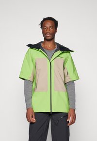 Cazadora corta de manga corta en verde y beige con capucha, que presenta un cierre frontal y un patrón de bloques de color contrastantes. Textura suave y corte relajado.